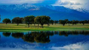 El tranquilo lago de Talayuela refleja los árboles y los picos nevados contra un cielo nublado, creando un entorno sereno para una Escapada Hotel Valles de Gredos + desayuno. - ForTwo - Planes para parejas