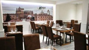 El moderno interior de un restaurante cuenta con sillas marrones, mesas de madera y un cautivador mural de un paisaje urbano. Cada mesa está adornada con un ramo de flores, lo que añade elegancia a este ambiente, inspirado en el Restaurante Bouquet (Cáceres). - ForTwo - Planes para parejas