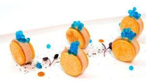 Sobre un plato blanco con guarniciones de colores, Restaurante Bouquet (Cáceres) presenta cinco galletas gourmet rellenas de cremoso relleno y artísticamente coronadas con esferas azules, ofreciendo un sabor de la elegancia cacereña en cada bocado. - ForTwo - Planes para parejas