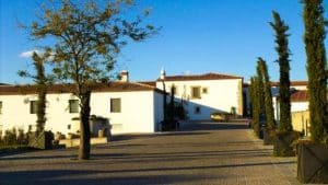 Edificios de estilo mediterráneo con tejados de teja se alzan bajo un cielo azul radiante, rodeados de cipreses. Cerca de allí, el elegante Restaurante Las Corchuelas Hospes Palacio de Arenales realza este idílico entorno con su encanto cacereño. - ForTwo - Planes para parejas