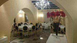 El elegante comedor del Restaurante Las Corchuelas Hospes Palacio de Arenales (Cáceres) cuenta con mesas redondas, sillas negras y doradas, lámparas de araña y un gran jarrón con flores rojas, que encapsulan el encanto de Cáceres y la sofisticación del lugar. - ForTwo - Planes para parejas