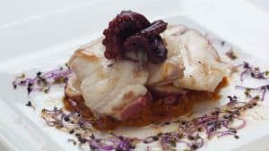 En el Restaurante Las Corchuelas Hospes Palacio de Arenales en Cáceres, saboree un plato bellamente presentado de pescado y pulpo a la parrilla en un plato blanco, adornado con brotes morados y hierbas. - ForTwo - Planes para parejas