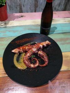 En el Restaurante Mario & Marieta de Cáceres, un plato negro con pulpo a la parrilla, salsa y especias reposa elegantemente junto a una botella alta en una colorida mesa de madera, capturando los vibrantes sabores locales. - ForTwo - Planes para parejas