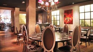 El restaurante Milo Grill en Barcelona ofrece un ambiente elegante con mesas redondas, sillas estampadas, una impresionante lámpara de araña y arte mural moderno. - ForTwo - Planes para parejas