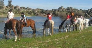 Los jinetes cruzan un río poco profundo con grandes rocas y extensiones de hierba, capturando la esencia de la Ruta a caballo por la dehesa extremeña (Cáceres). - ForTwo - Planes para parejas