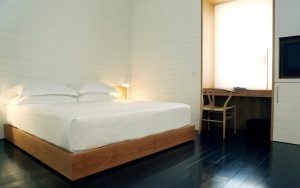 La habitación minimalista del Hotel Atrio de Cáceres incluye una cama de madera, sábanas blancas impecables y un elegante escritorio con silla. Una lámpara junto al televisor proporciona una iluminación suave, todo ello complementado por un elegante suelo de madera oscura. - ForTwo - Planes para parejas