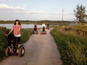 La gente se desliza en la Ruta en Segway por los arrozales de Pals a lo largo de un camino de grava junto a un lago, rodeado de exuberante vegetación y árboles imponentes, disfrutando de un paseo sereno por la naturaleza. - ForTwo - Planes para parejas