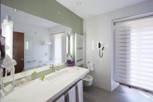 Un baño moderno que evoca vibraciones de viajes de lujo como El Parador de Cáceres, con un gran espejo, encimera blanca, toallas de felpa y una ventana con persianas horizontales. - ForTwo - Planes para parejas