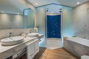 Descubra un moderno baño en el Hotel Barceló V Centenario, Cáceres, con lavabos dobles, encimera de mármol, una lujosa bañera y una ducha de cristal resaltada por elegantes azulejos geométricos azules. - ForTwo - Planes para parejas