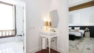Un baño blanco con lavabo y espejo conduce a un lujoso dormitorio con una cama grande y balcón, capturando la tranquilidad de la "Escapada de lujo en el hotel Palacio de los Patos con spa incluido (Granada)" para la máxima relajación. - ForTwo - Planes para parejas