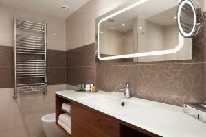 El sofisticado baño del Hotel Hilton de Barcelona cuenta con un amplio espejo, un lavabo moderno y artículos de aseo organizados. Un toallero realza las elegantes paredes de azulejos, ofreciendo una experiencia de lujo. - ForTwo - Planes para parejas