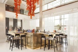 Un bar contemporáneo en el Hotel Hilton (Barcelona) cuenta con taburetes altos, una barra central y una llamativa obra de arte en rojo. Grandes ventanales inundan el espacio de luz natural, capturando el ambiente animado de Barcelona. - ForTwo - Planes para parejas