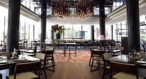 El Hilton Barcelona cuenta con un moderno interior de restaurante con columnas oscuras, una mesa central, iluminación decorativa y grandes ventanales que ofrecen una experiencia gastronómica con estilo. - ForTwo - Planes para parejas