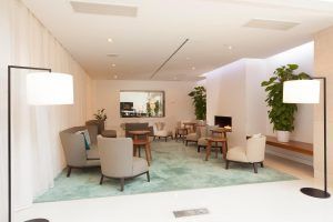 Disfrute del moderno salón del Hotel Hilton (Barcelona), con sillones y mesitas sobre una exuberante alfombra verde. Cálidamente iluminado por lámparas de pie, el espacio luce paredes blancas, techo y plantas vibrantes junto a una chimenea crepitante. - ForTwo - Planes para parejas