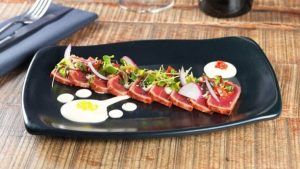 El atún sellado en rodajas sobre un plato negro, adornado con verduras, rábanos y gotas de salsa, encarna la esencia culinaria de Cáceres en las encantadoras mesas de madera del Restaurante Mastropiero Gastrobar. - ForTwo - Planes para parejas