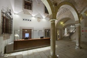 El imponente vestíbulo del Parador de Cáceres exhibe una amplia elegancia con sus columnas de piedra arqueadas, que evocan la arquitectura de la ciudad. Con un acogedor mostrador de recepción y una imponente escalera, captura el encanto atemporal de su homónimo. - ForTwo - Planes para parejas