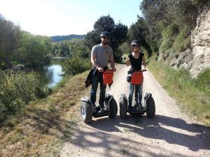 Dos personas en la "Ruta en Segway en los arrozales de Pals" recorren un camino de tierra junto a un río, con casco y gafas de sol, rodeados de frondosos árboles y arrozales cercanos. - ForTwo - Planes para parejas