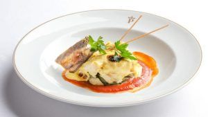 Este plato gourmet del Restaurante Torreorgaz Parador de Cáceres presenta pescado preparado por expertos, puré de papas cremoso y una guarnición vibrante en un plato blanco con el logotipo decorativo del restaurante. - ForTwo - Planes para parejas