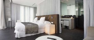 Disfrute de una escapada de lujo en el Hotel Palacio de los Patos, Granada, que cuenta con una moderna habitación con acceso al spa, una cama doble con sábanas blancas, una elegante cabecera de madera y un atractivo baño tipo spa parcialmente visible. - ForTwo - Planes para parejas