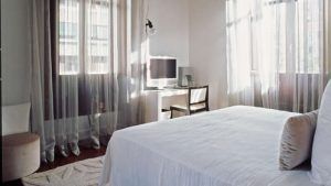 Disfrute de una estancia de lujo en el Hotel Palacio de los Patos en una luminosa habitación con cortinas transparentes, cama blanca, escritorio de madera, silla y televisor de pared. Disfrute del diseño moderno y relájese en el exquisito spa para una experiencia inigualable con Escapada de lujo en el hotel Palacio de los Patos con spa incluido (Granada). - ForTwo - Planes para parejas