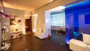 Experimente el lujo moderno en el spa del Hotel Palacio de los Patos en Granada, con una piscina iluminada con luz azul, elegantes asientos blancos, suelos de madera y estanterías decoradas con gusto. Esta Escapada de lujo ofrece una experiencia de spa inigualable. - ForTwo - Planes para parejas