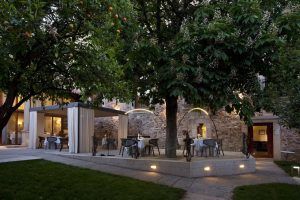 Un comedor al aire libre con mesas, sillas y un toldo iluminado bajo un árbol con un edificio de piedra como telón de fondo evoca el encantador ambiente del Restaurante Torreorgaz Parador de Cáceres. - ForTwo - Planes para parejas