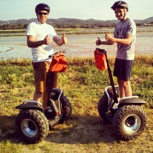 Dos personas con cascos se deslizan por los pintorescos campos de arrozales de Pals en una "Ruta en Segway en los arrozales de Pals", haciendo gestos de aprobación con los pulgares hacia arriba mientras señalan las extensiones de hierba y las suaves colinas. - ForTwo - Planes para parejas