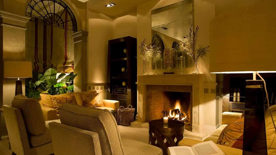 Una acogedora sala de estar con chimenea, exuberantes plantas e iluminación ambiental evoca el ambiente del Hotel Villa Oniria. Equipada con un cómodo sofá y sillones, es ideal para una escapada romántica, al igual que la "Escapada romántica con spa en el hotel Villa Oniria 4 estrellas (Granada)". - ForTwo - Planes para parejas