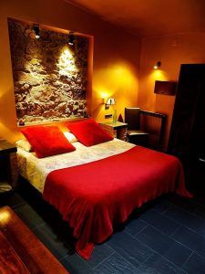 El acogedor dormitorio de la Almazara de San Pedro cuenta con una cálida iluminación, una rústica pared de piedra y una colcha roja con almohadas a juego en la cama doble. Esta encantadora casa rural se encuentra cerca de la Sierra de Gata, en Cáceres, e incluye un baño árabe. - ForTwo - Planes para parejas