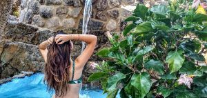 Una persona bajo una cascada entre rocas y flores, que recuerda a una escapada serena como la Almazara de San Pedro - Casa rural en Sierra de Gata con baño árabe (Cáceres), captura la belleza natural escondida. - ForTwo - Planes para parejas