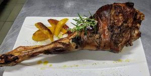 Pierna de cordero asada con hierbas aromáticas en plato, evocando el ambiente de la Almazara de San Pedro - Casa rural en Sierra de Gata, adornada con patatas en rodajas y romero. - ForTwo - Planes para parejas