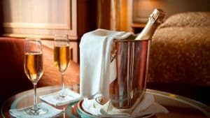 Una botella de cava se enfría en una cubitera con hielo cubierta con un paño blanco, rodeada de dos copas llenas sobre una mesa de cristal, creando un tête-à-tête perfecto que recuerda a una estancia en el Escapada Hotel SixtyTwo de la vibrante Barcelona. - ForTwo - Planes para parejas