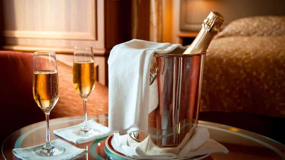Una botella de cava se enfría en una cubitera con hielo cubierta con un paño blanco, rodeada de dos copas llenas sobre una mesa de cristal, creando un tête-à-tête perfecto que recuerda a una estancia en el Escapada Hotel SixtyTwo de la vibrante Barcelona. - ForTwo - Planes para parejas