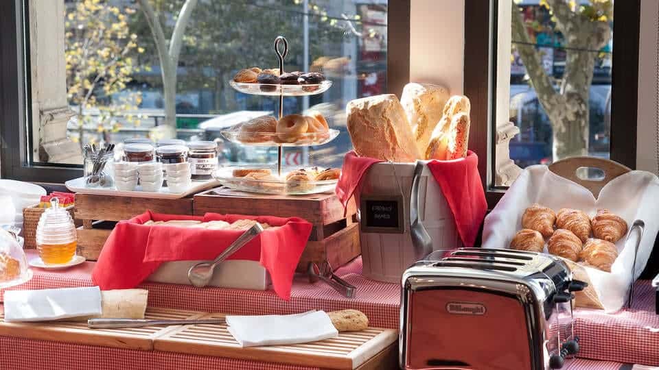 Disfrute de un delicioso desayuno buffet en el Escapada Hotel SixtyTwo con bollería, pan, fruta en brochetas y mermelada sobre un mantel rojo a cuadros junto a la ventana. Añada un toque de cava a su mañana para empezar el día de la mejor manera en Barcelona. - ForTwo - Planes para parejas