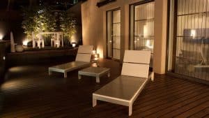 Relájese en un moderno patio exterior con dos tumbonas y una mesa, rodeado de exuberante vegetación y una cálida iluminación en una terraza de madera. Disfrute de un cava que evoca la tranquilidad del Escapada Hotel SixtyTwo. - ForTwo - Planes para parejas