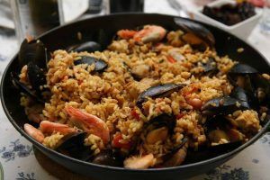 Una paella con mejillones, gambas y arroz reposa sobre un mantel floral, que evoca el casco histórico de Valencia. Aquí, el Escapada Hotel Zenit ofrece a los aficionados a la gastronomía una visita con paella, acceso a la sauna, gimnasio y visitas guiadas. - ForTwo - Planes para parejas