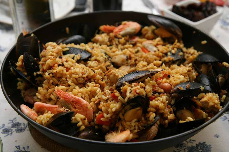 Una paella con mejillones, gambas y arroz reposa sobre un mantel floral, que evoca el casco histórico de Valencia. Aquí, el Escapada Hotel Zenit ofrece a los aficionados a la gastronomía una visita con paella, acceso a la sauna, gimnasio y visitas guiadas. - ForTwo - Planes para parejas