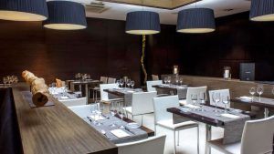 El moderno restaurante en el casco histórico de Valencia presume de paredes de madera oscura, sillas blancas, mesas elegantemente dispuestas, grandes lámparas colgantes y un tronco decorativo. Es un espacio acogedor que realza el ambiente refinado del Hotel Zenit, como parte de la experiencia "Escapada". - ForTwo - Planes para parejas