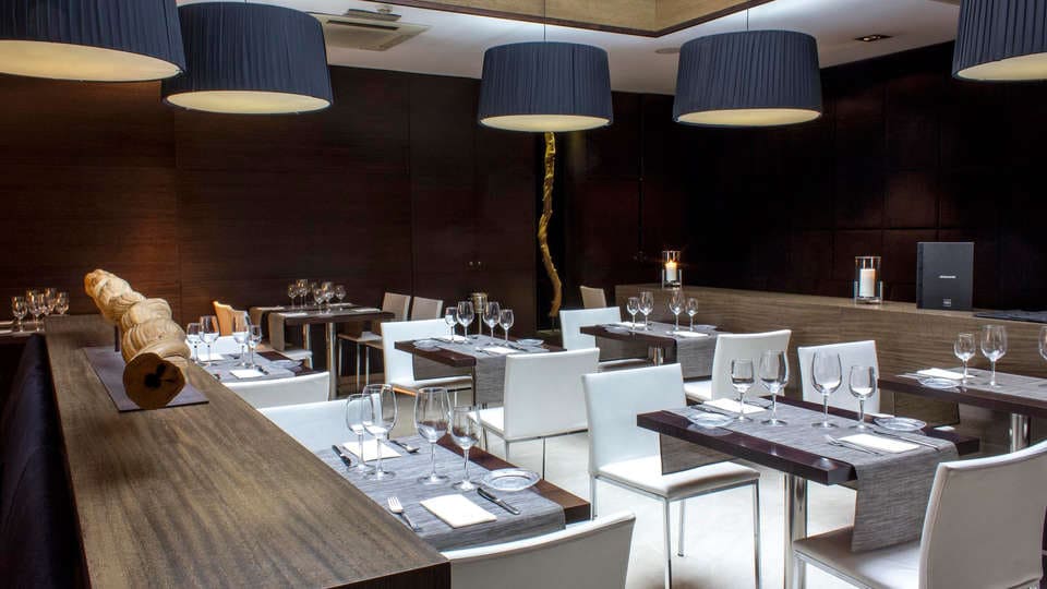 El moderno restaurante en el casco histórico de Valencia presume de paredes de madera oscura, sillas blancas, mesas elegantemente dispuestas, grandes lámparas colgantes y un tronco decorativo. Es un espacio acogedor que realza el ambiente refinado del Hotel Zenit, como parte de la experiencia "Escapada". - ForTwo - Planes para parejas