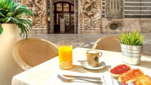 Disfrute de una "Escapada SH Inglés Boutique + desayuno (Valencia)" con una mesa de cafetería al aire libre con zumo de naranja, café, tostadas y vegetación frente al ornamentado telón de fondo arquitectónico de SH Inglés Boutique. - ForTwo - Planes para parejas