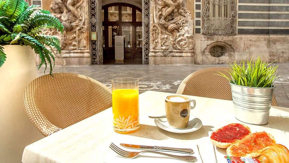 Disfrute de una "Escapada SH Inglés Boutique + desayuno (Valencia)" con una mesa de cafetería al aire libre con zumo de naranja, café, tostadas y vegetación frente al ornamentado telón de fondo arquitectónico de SH Inglés Boutique. - ForTwo - Planes para parejas