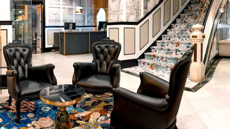 Experimente el elegante vestíbulo de Escapada SH Inglés Boutique + desayuno (Valencia), con tres sillas negras, una alfombra floral y una escalera ornamentada. Perfecto para su próxima escapada o para disfrutar de un desayuno relajado con estilo. - ForTwo - Planes para parejas