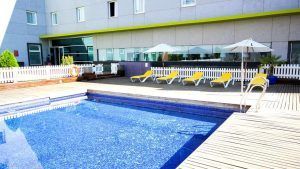 La piscina al aire libre de azulejos azules se ve reforzada por una terraza de madera y tumbonas de color amarillo vibrante bajo sombrillas blancas, lo que proporciona un lugar ideal para relajarse después de visitar sitios cercanos como el Gran Casino Aljarafe, como parte de la Escapada con desayuno, bañera de hidromasaje y entradas al Gran Casino Aljarafe. - ForTwo - Planes para parejas