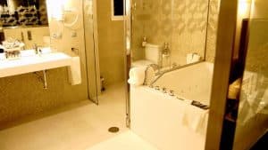 Transforme su baño en un refugio que recuerde a la Escapada con piscina, cocktel y visita guiada (Sevilla) con un moderno lavabo blanco, ducha de cristal, inodoro y una gran bañera contra paredes de azulejos bajo una iluminación cálida. - ForTwo - Planes para parejas