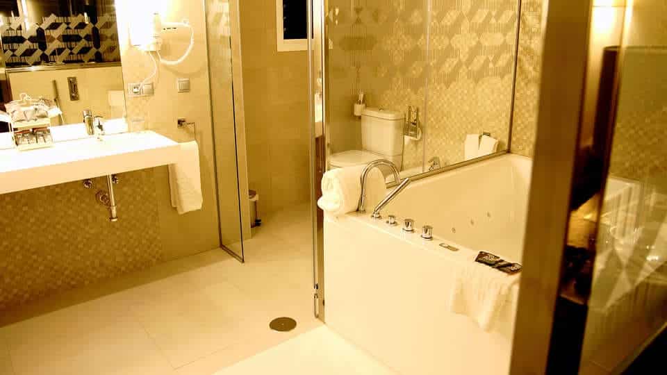 Transforme su baño en un refugio que recuerde a la Escapada con piscina, cocktel y visita guiada (Sevilla) con un moderno lavabo blanco, ducha de cristal, inodoro y una gran bañera contra paredes de azulejos bajo una iluminación cálida. - ForTwo - Planes para parejas