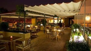Disfrute de una velada encantadora en este restaurante al aire libre, donde las mesas y sillas de madera y un toldo de tela crean un ambiente acogedor. La iluminación tenue y las plantas realzan el ambiente, convirtiéndolo en el lugar perfecto para disfrutar de una escapada con piscina, cóctel y visita guiada bajo las estrellas en Sevilla. - ForTwo - Planes para parejas