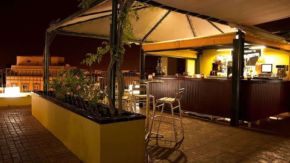 Por la noche, disfrute de la "Escapada con piscina, cóctel y visita guiada (Sevilla)" en el bar de la azotea con un mostrador iluminado, taburetes y jardineras llenas de flores mientras saborea un cóctel artesanal y contempla la vista. - ForTwo - Planes para parejas