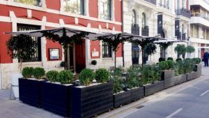 Un café callejero con grandes sombrillas y plantas en macetas afuera de un edificio rojo con ventanas ornamentadas captura el estilo encantador de Valencia, que recuerda al área cerca del Restaurante Ampar en Palau de la Mar. - ForTwo - Planes para parejas