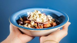 Unas manos que sostienen un bol azul de ensalada griega con queso feta, aceitunas y tomates sobre un fondo azul evocan los sabores del Restaurante griego Kuzina (Valencia). - ForTwo - Planes para parejas