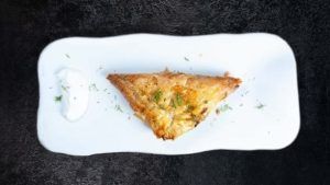Un pastel triangular con hierbas y crema adorna un plato blanco rectangular en el Restaurante griego Kuzina en Valencia, enmarcado sobre un fondo oscuro. - ForTwo - Planes para parejas